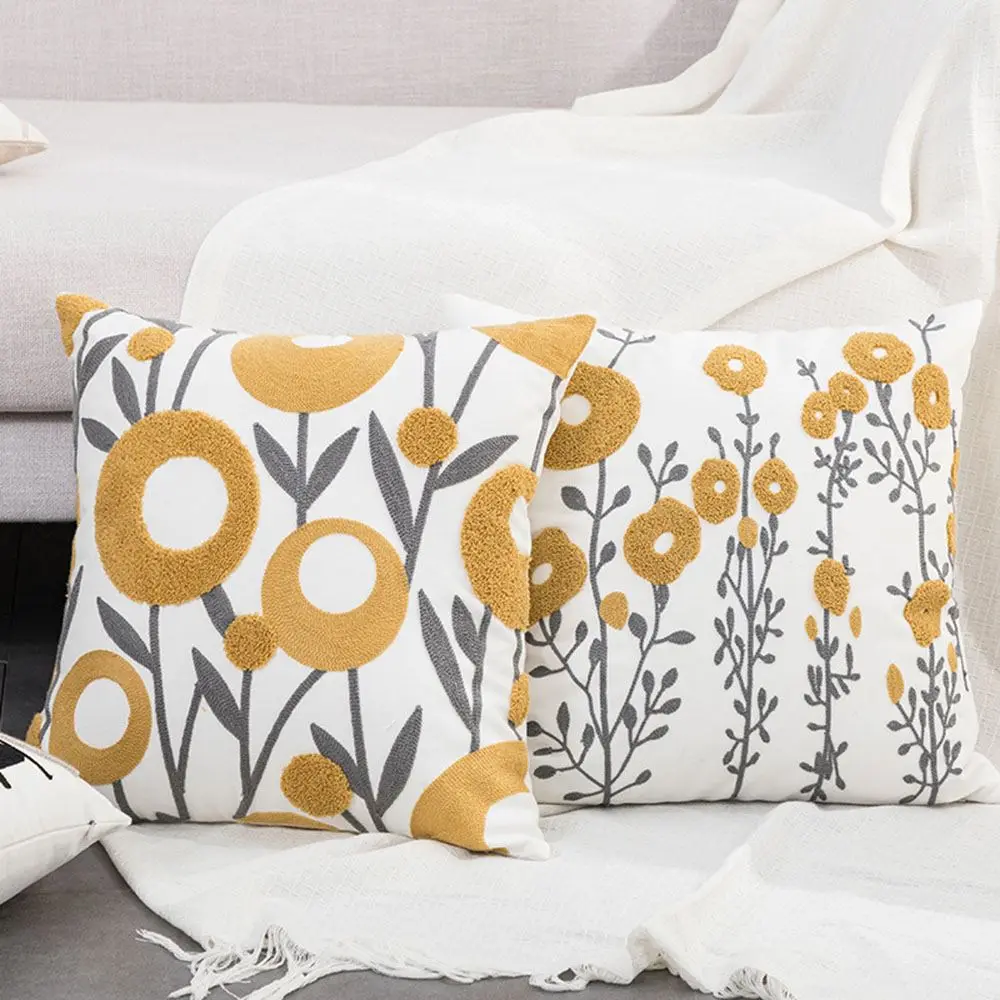 Zwei Kissenhüllen mit senfgelben gestickten Blumen und grauen Ranken auf einem hellen Sofa
